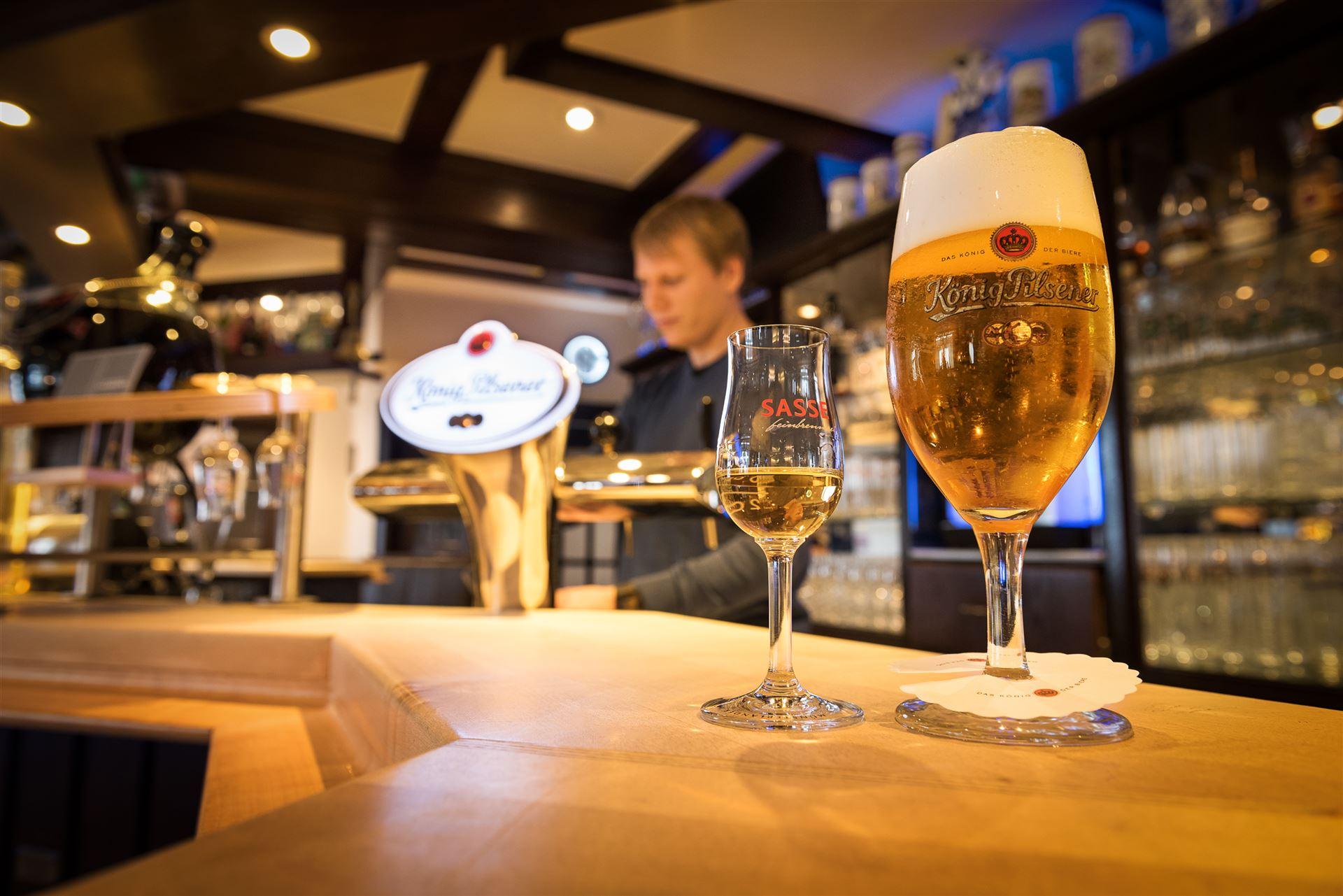 Gedeelte van de verlichte bar met op de voorgrond een glas könig pilsener en een Sasse likeur.